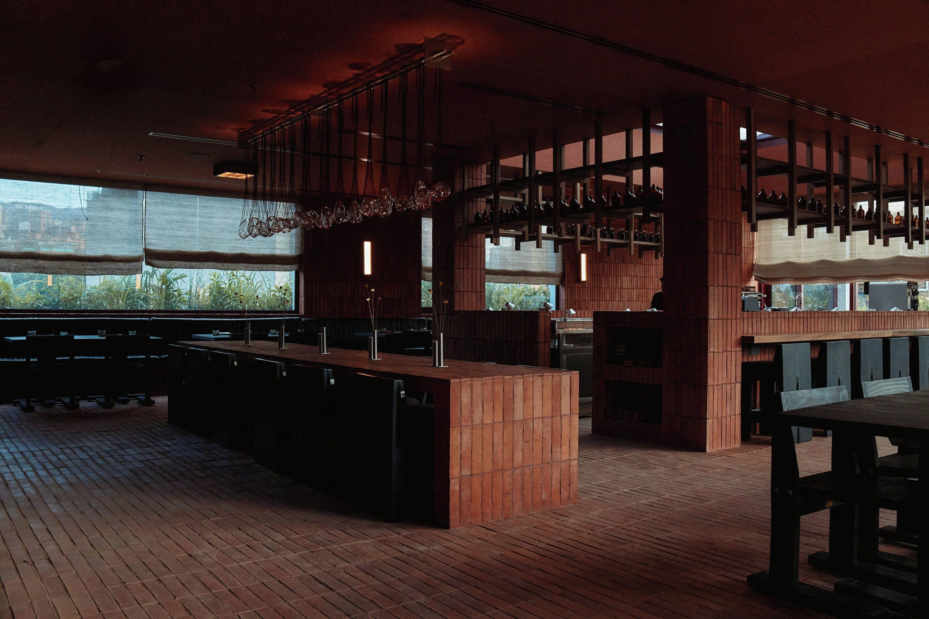 Un interior de restaurante con suelo de ladrillo, mesas y bancos de madera, lámparas colgantes y ventanas amplias.