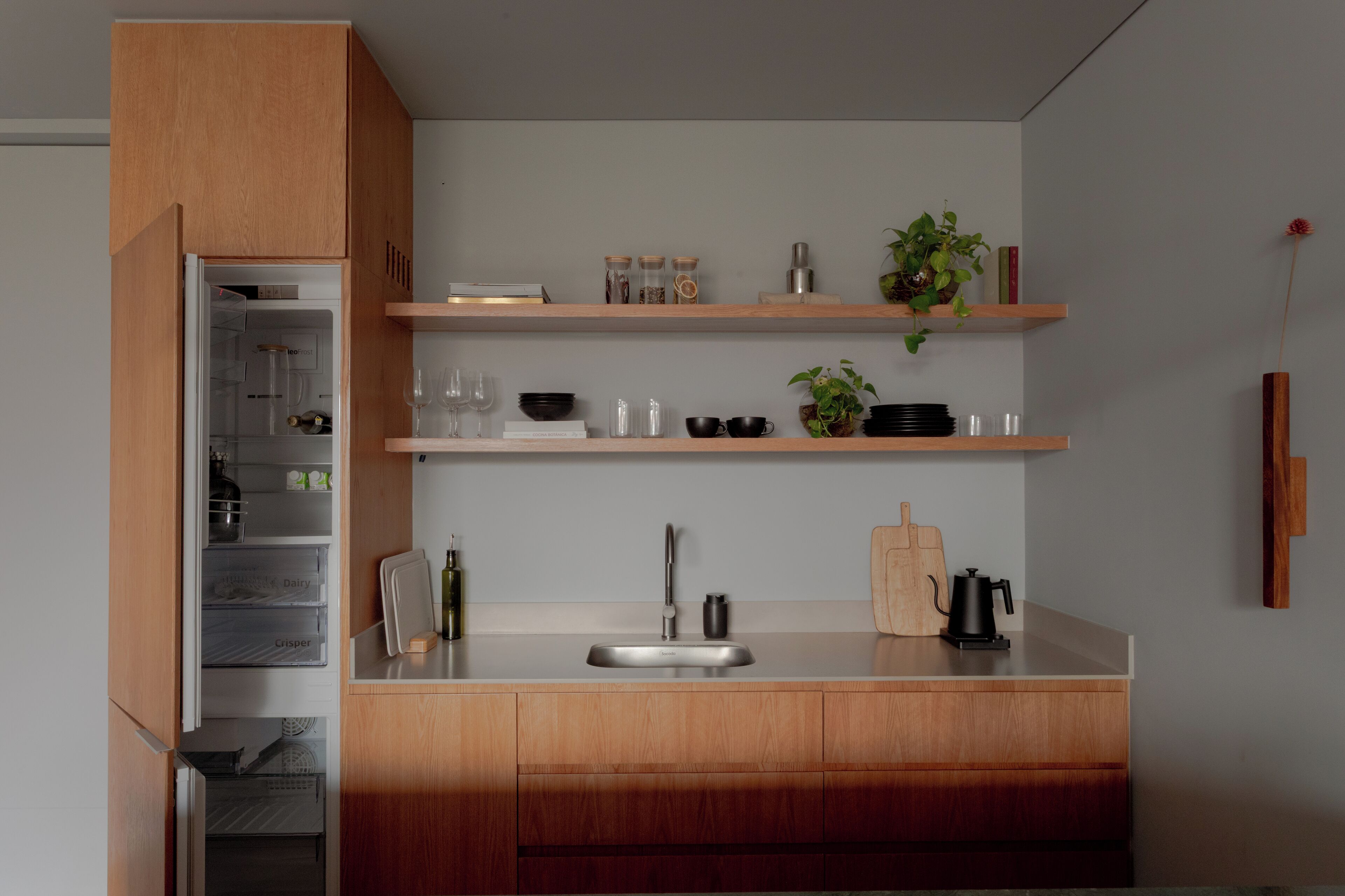 A modern kitchen with wooden cabinets, a stainless steel sink, and open shelving with kitchenware and plants.