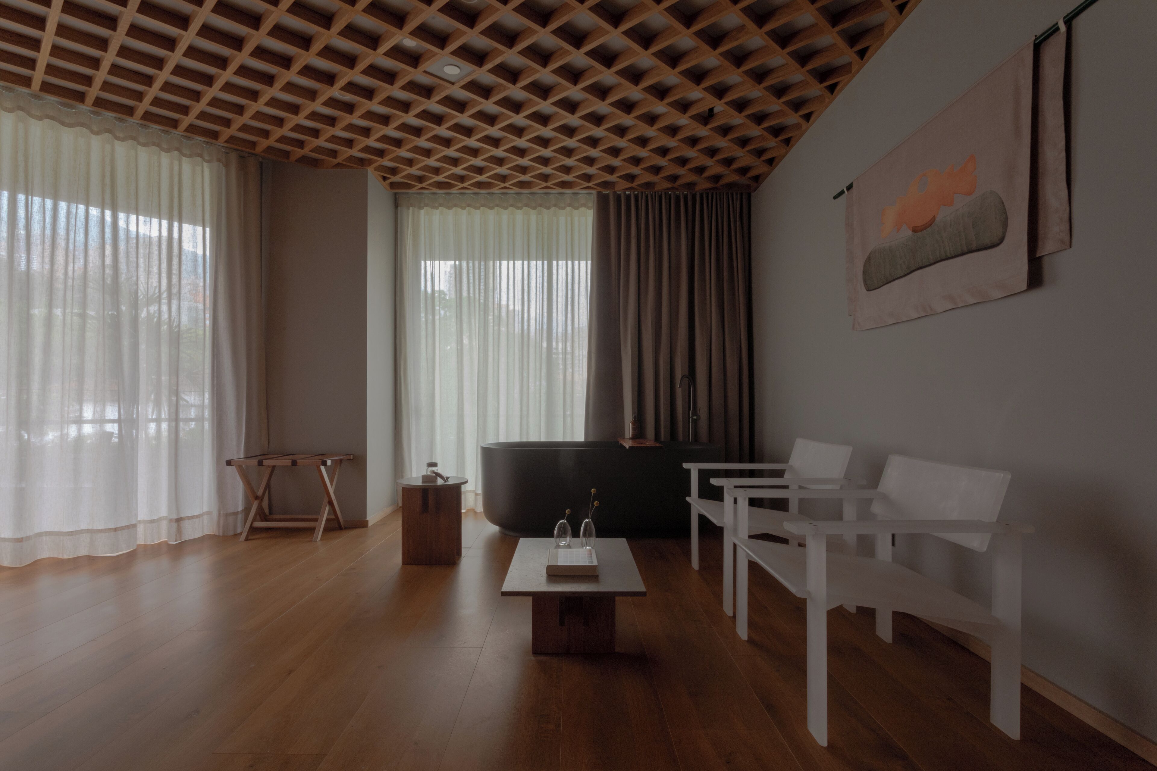 A modern living room with a wooden ceiling, a large window with sheer curtains, a dark sofa, white chairs, and a small table with decorative items.