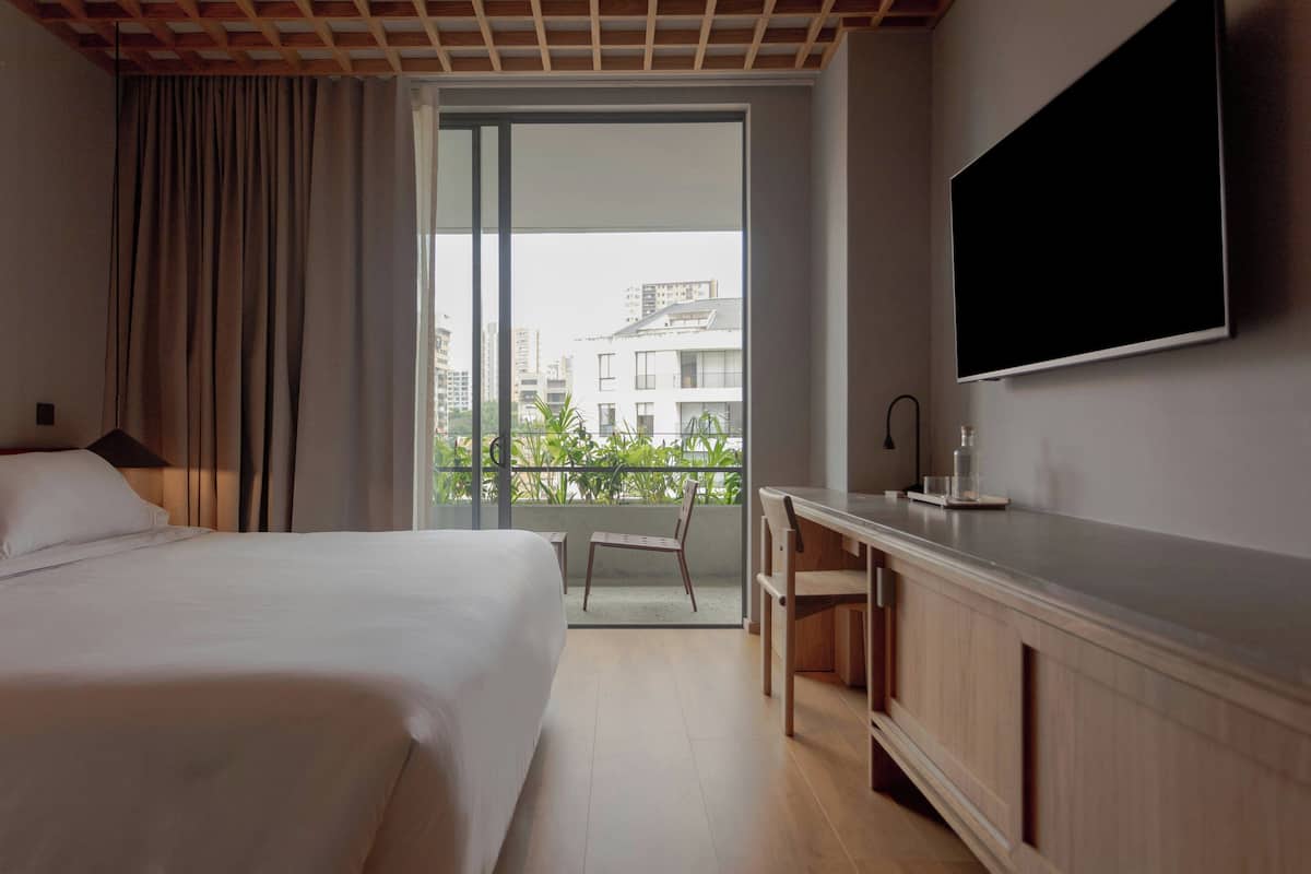 A hotel room with a bed, a desk, a chair, a TV, and a balcony with a view of buildings.