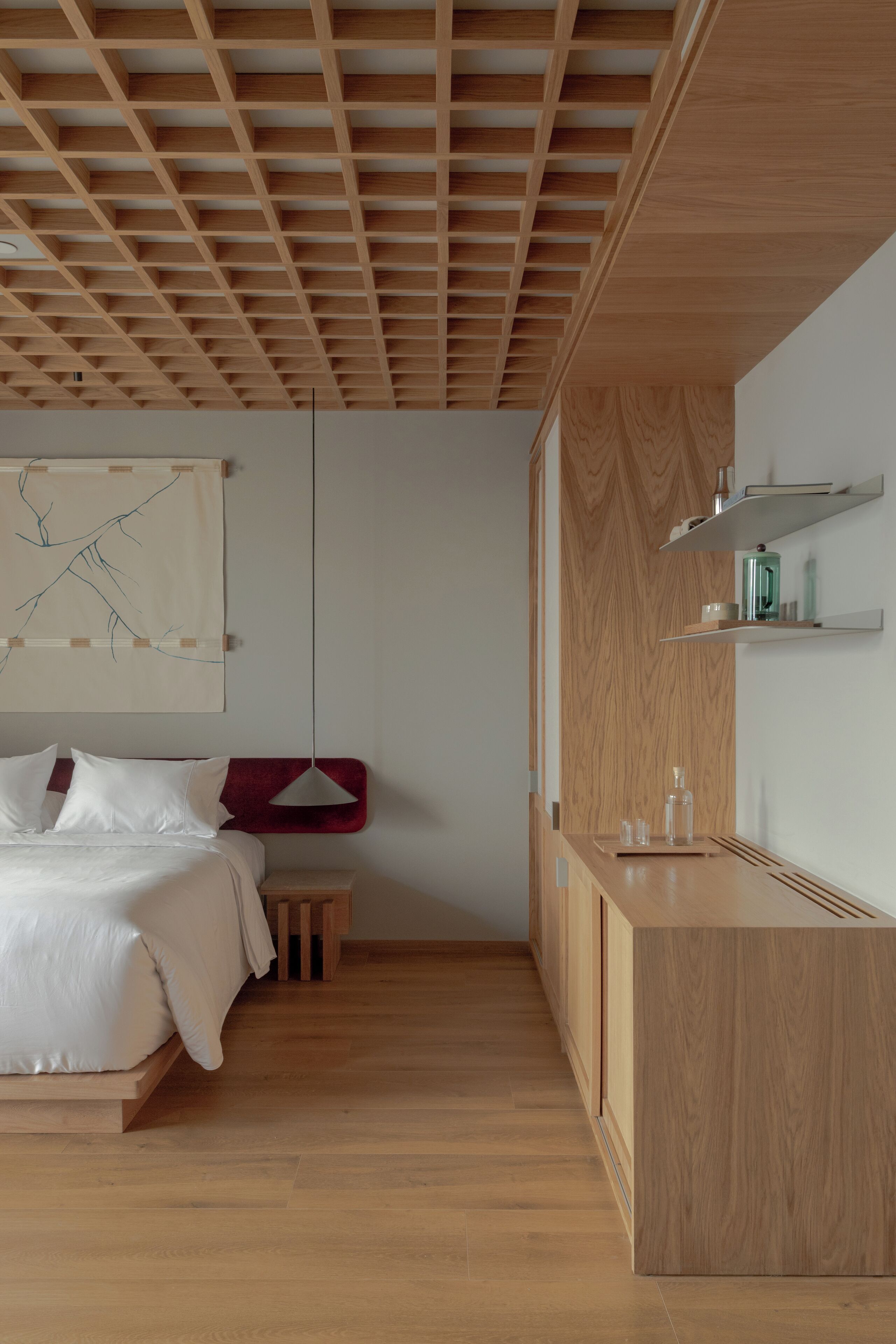 A modern bedroom with a wooden ceiling, a bed with white bedding, a small bedside table, and a wooden cabinet with shelves.