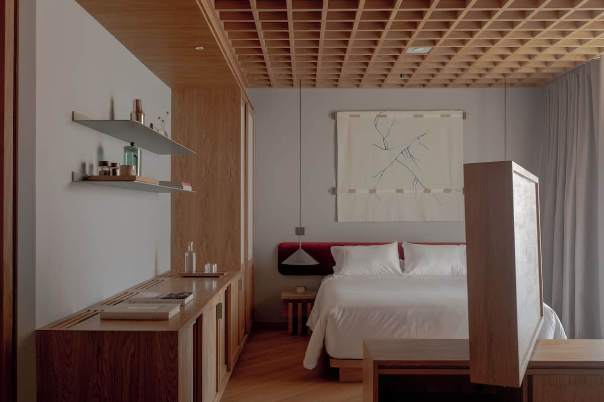 A modern hotel room with a wooden ceiling, a bed with white linens, a bedside table, a small nightstand, and a wall-mounted shelf with decorative items.