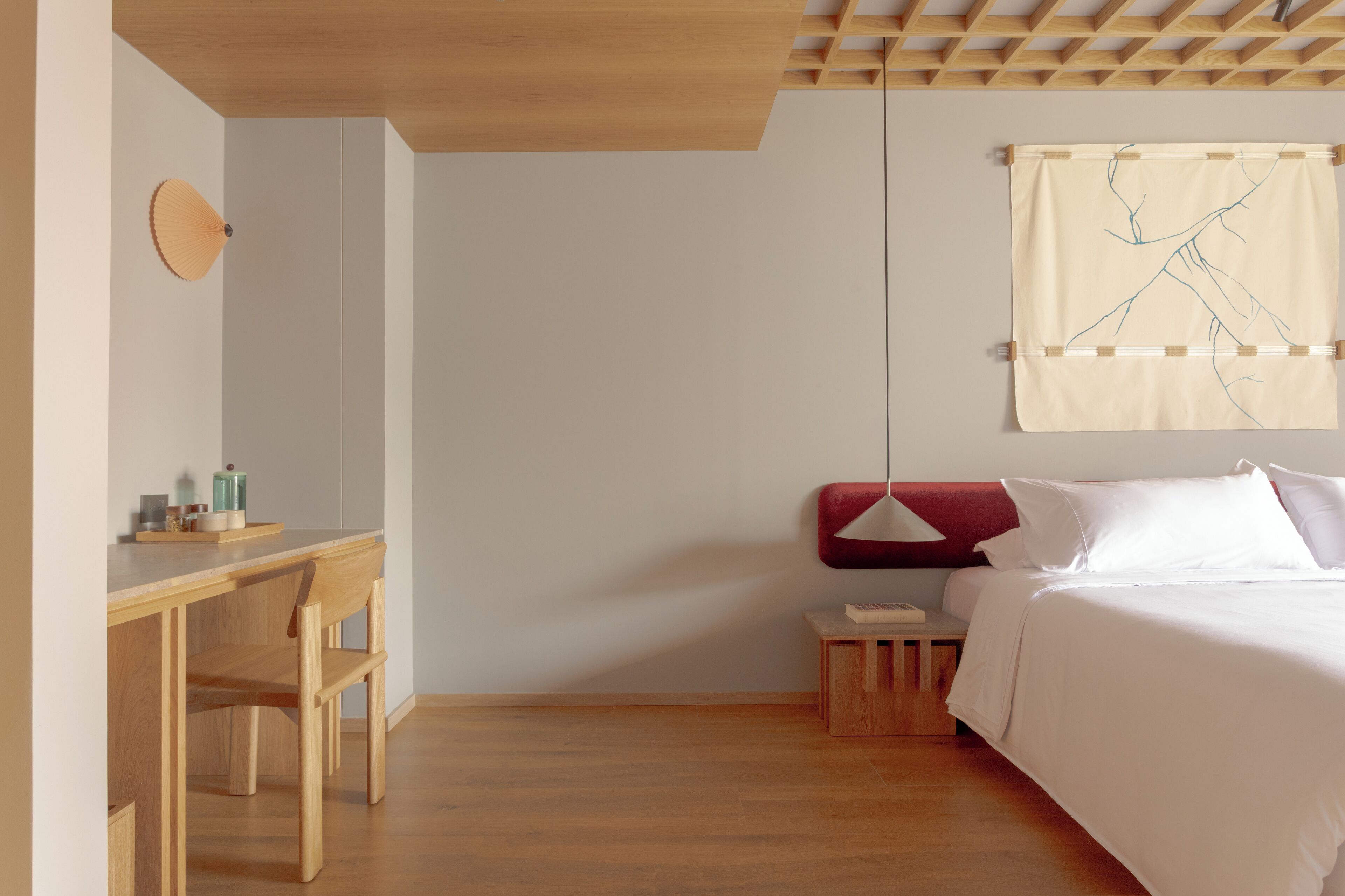 Un dormitorio minimalista con un escritorio de madera, una cama con ropa de cama blanca y un tapiz colgante.