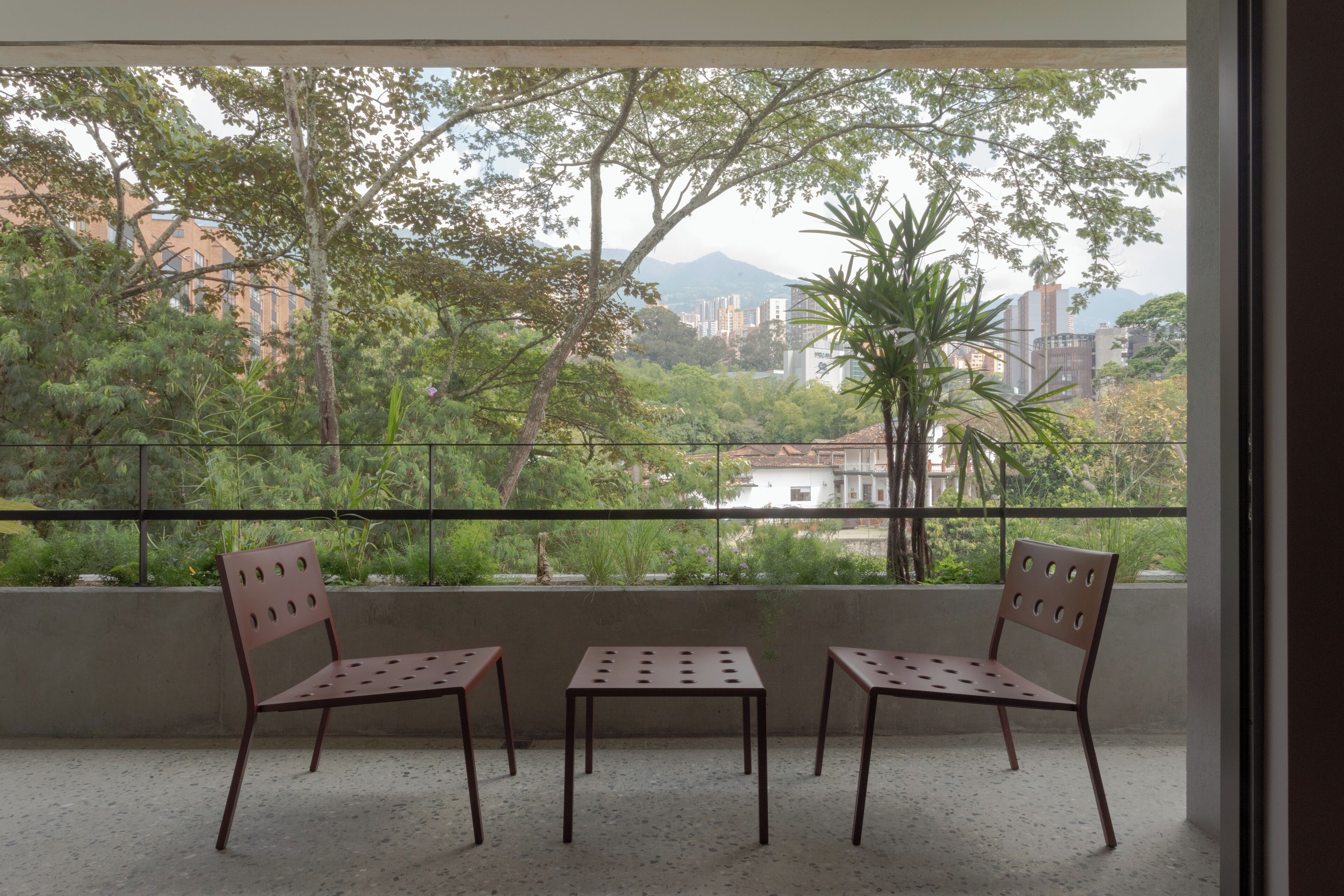 Un balcon avec deux chaises et une table, d’où l’on peut admirer une vue sur la ville, avec ses bâtiments et ses arbres.