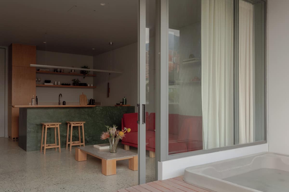 A modern living room with a glass door, a red sofa, a wooden coffee table, and a kitchen area with wooden shelves and a marble countertop.