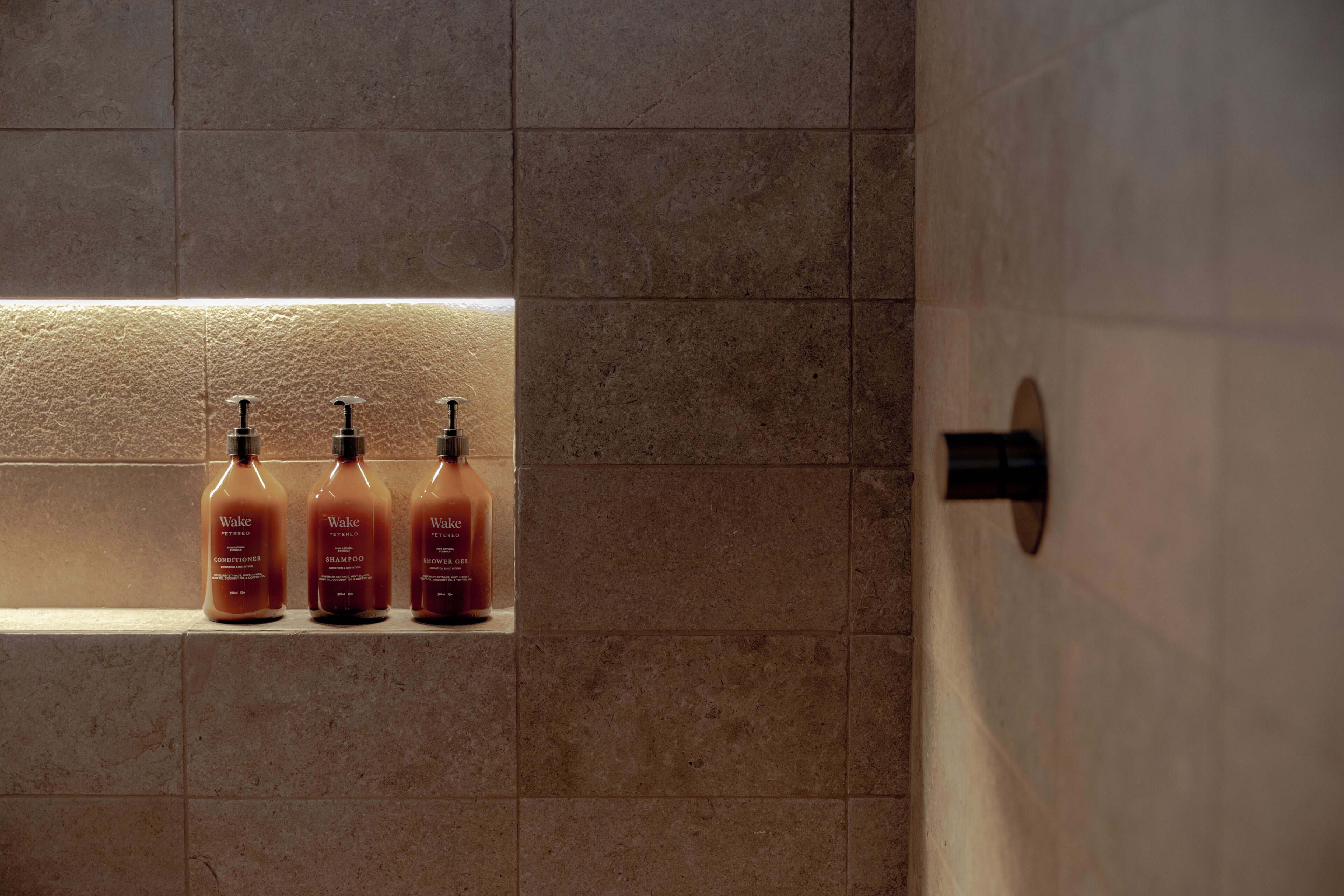 Three Wake brand toiletries in a bathroom with tiled walls and a built-in shelf.