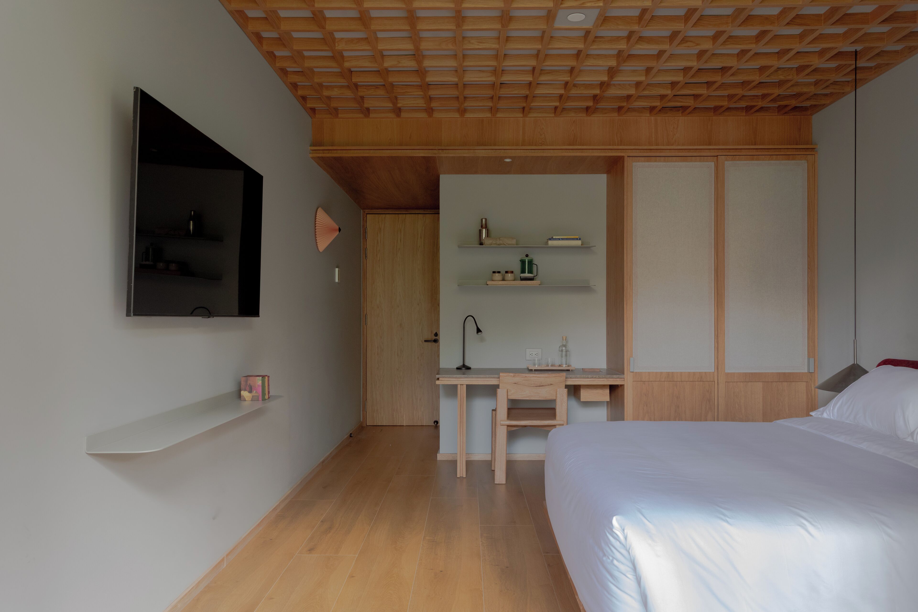 A modern bedroom with a wooden ceiling, a bed, a desk with a chair, a TV mounted on the wall, and a wardrobe with sliding doors.