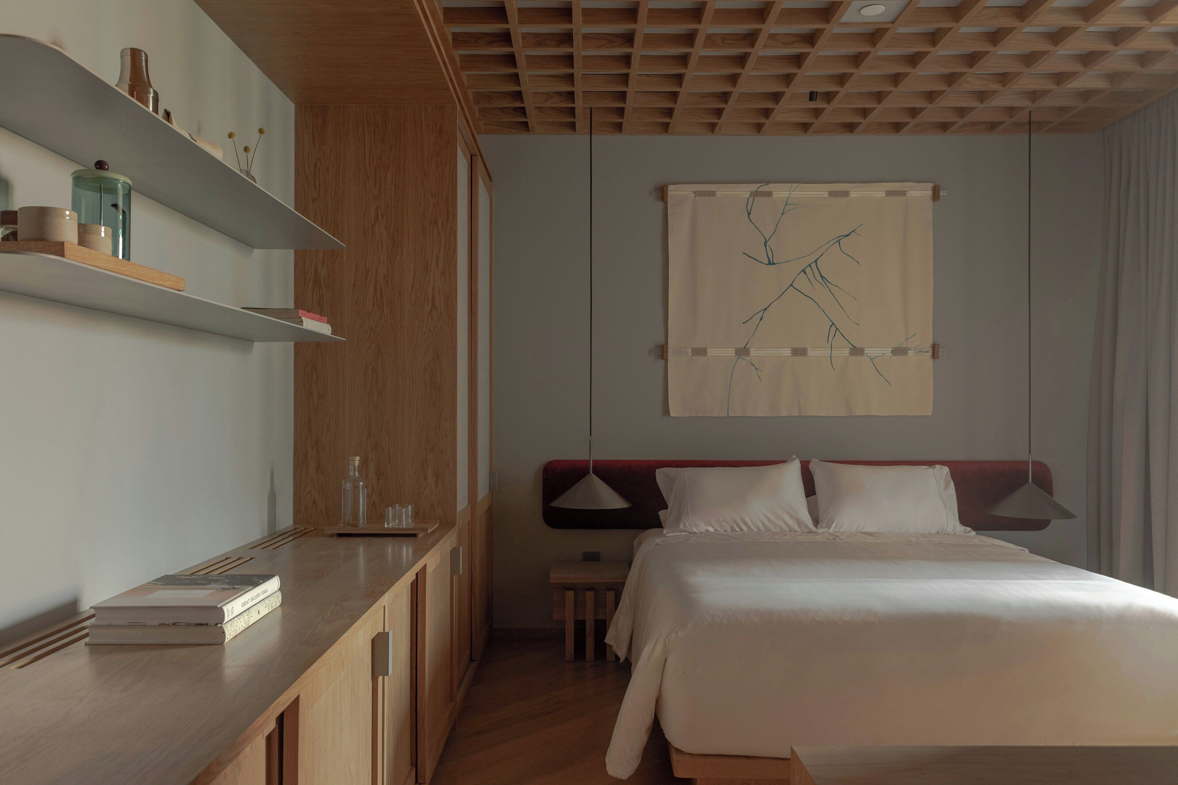 Una habitación de hotel con un escritorio de madera, una cama con ropa de cama blanca, una obra de arte abstracta colgada en la pared y un techo con diseño de entramado de madera.