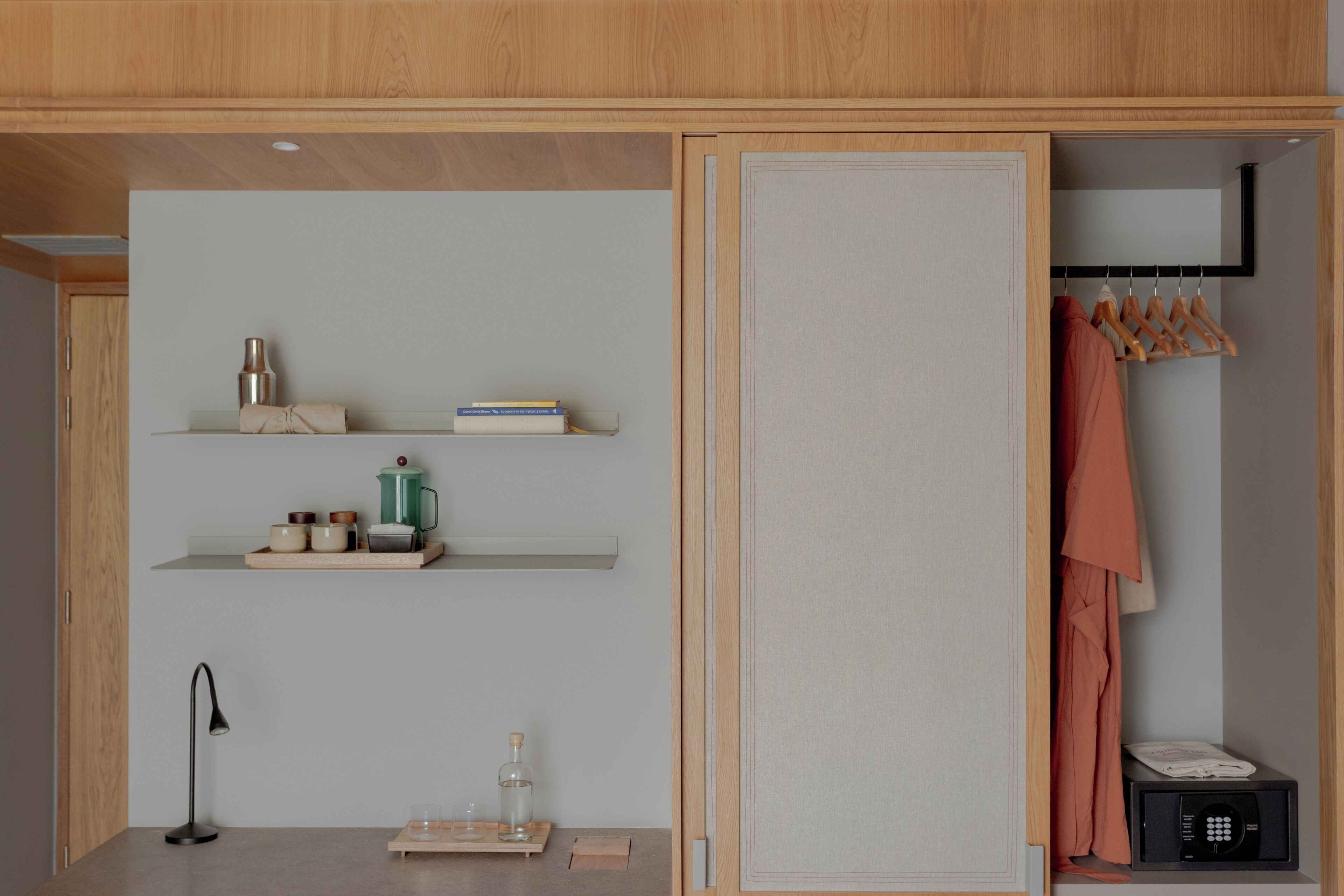 A modern room with wooden paneling, a wardrobe with hanging clothes, shelves with books and decorative items, and a small table with a lamp and glassware.