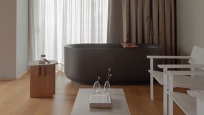 A modern living room with a wooden ceiling, a large window with sheer curtains, a dark sofa, a white table, and a small wooden side table. - Wake Medellin, a Member of Design Hotels (Medellín)