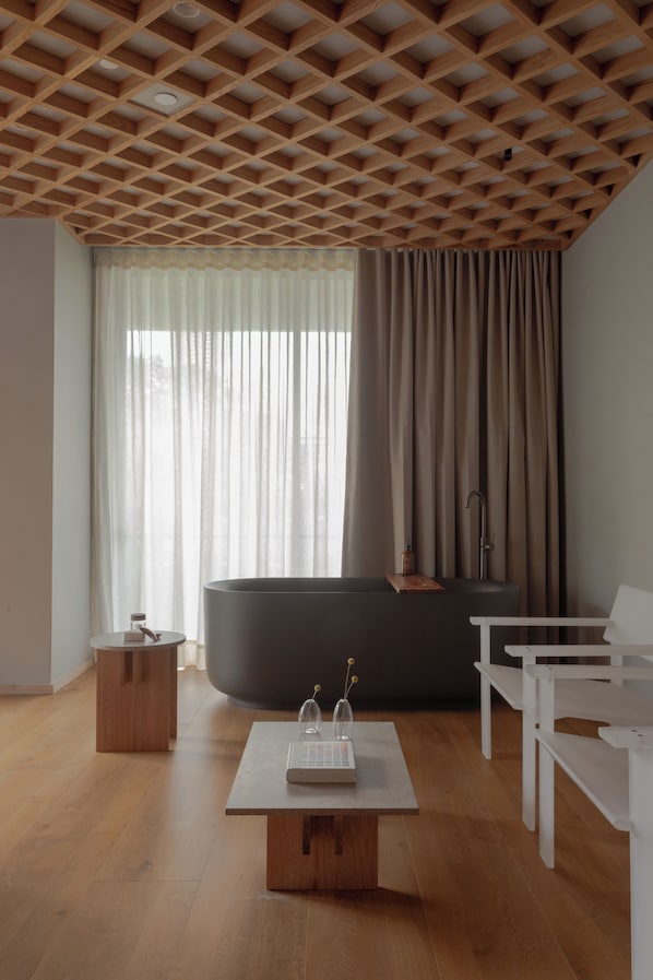 A modern living room with a wooden ceiling, a large window with sheer curtains, a dark sofa, a white table, and a small wooden side table. - Wake Medellin, a Member of Design Hotels (Medellín)
