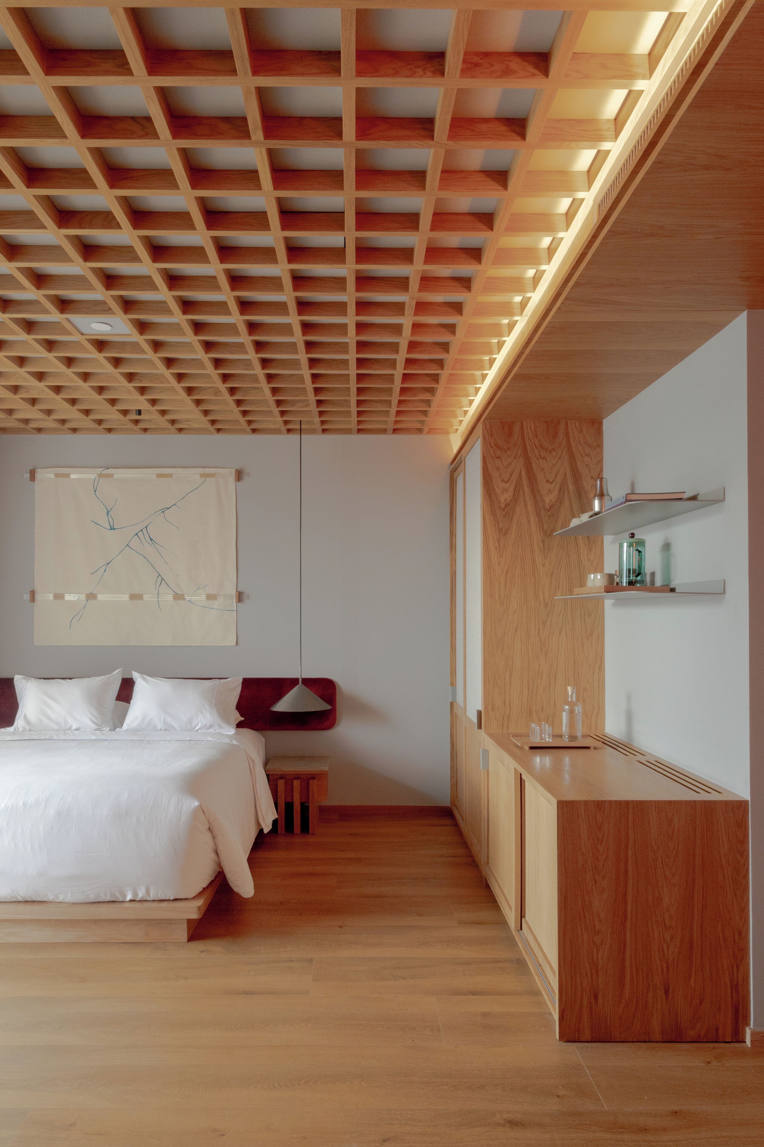 Ein modernes Schlafzimmer mit Holzbalkendecke, einem Bett mit weißen Leinentüchern, einem Nachttisch und einem an der Wand befestigten Ablagefach mit Pflegeutensilien.