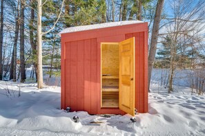 Een rode houten tuinschuur met een open deur, gelegen in een besneeuwd bos met een meer op de achtergrond.