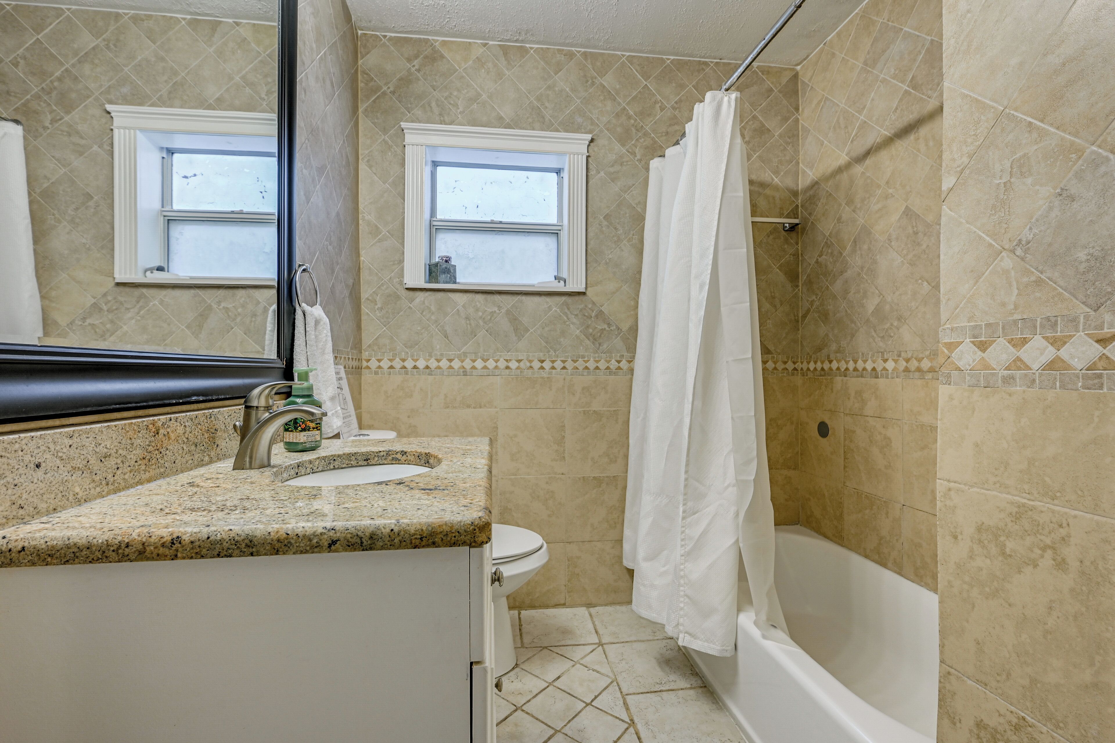 A bathroom with a granite countertop, a white toilet, a bathtub, and two windows.