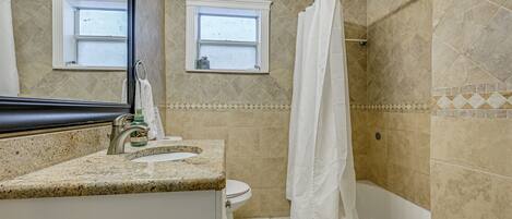 A bathroom with a granite countertop, a white toilet, a bathtub, and two windows.