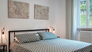 Ein Schlafzimmer mit einem Bett, zwei an der Wand befestigten Kunstwerken, einem Nachttisch mit Lampe und einem Fenster mit Blick auf Grünflächen.
