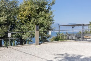 Ein Kiesplatz mit Metallzaun, einem Technikschacht und einer überdachten Sitzgelegenheit mit Blick auf ein Gewässer und Berge in der Ferne.