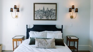 A bed with a dark headboard, white bedding, and multiple pillows. There are two nightstands with lamps on either side of the bed. A framed cityscape artwork is centered above the bed.
