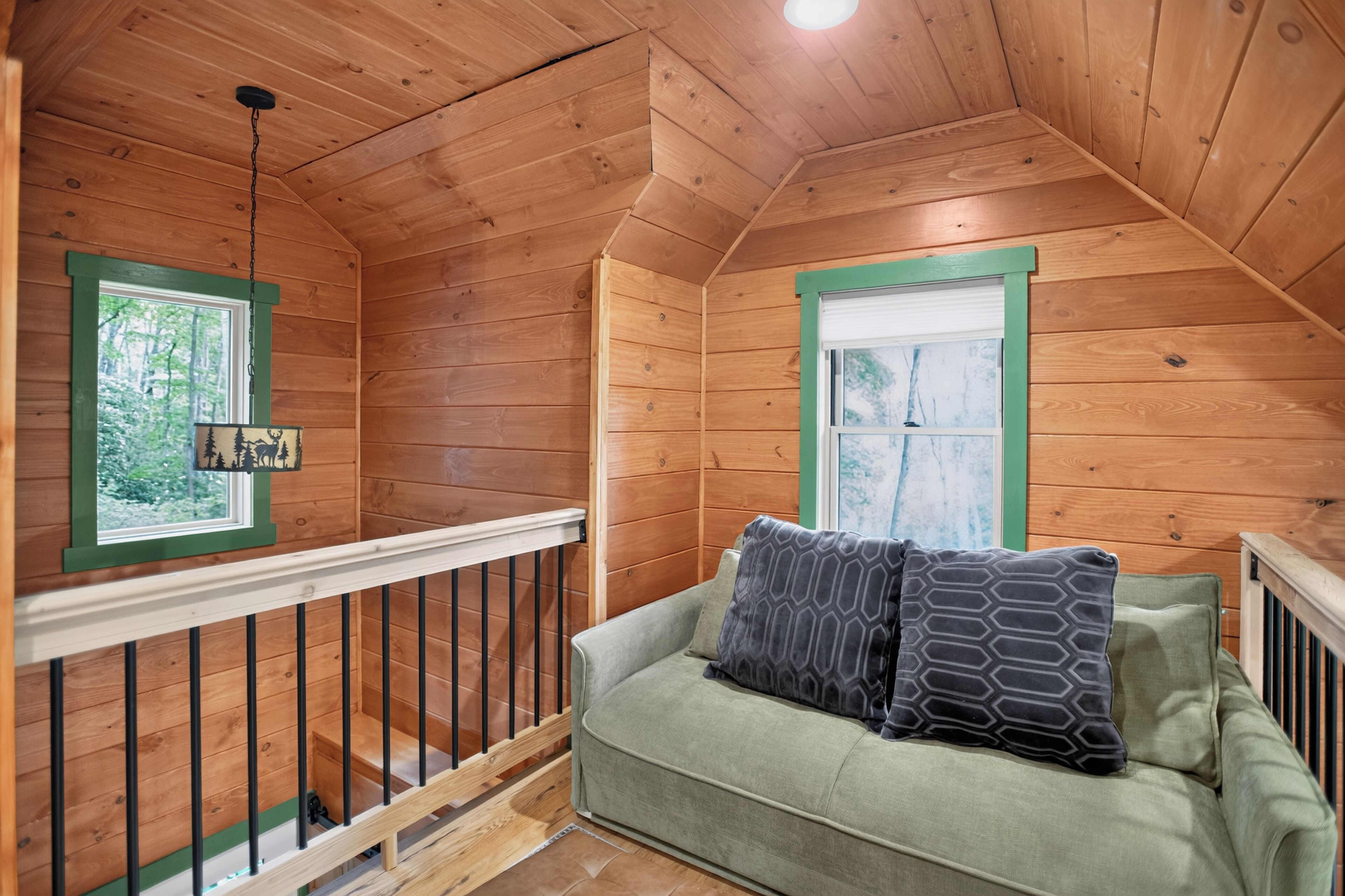 A wooden cabin interior with a green couch, two patterned pillows, a window with a view of trees, and a hanging light fixture.