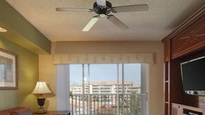 Sala de estar com sofá de couro marrom, mesa de centro de madeira, ventilador de teto, TV sobre um móvel de madeira e varanda com vista para prédios.