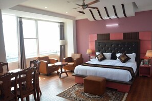A hotel room with a large bed, a ceiling fan, a dining area with chairs and a table, and a view of the outdoors through large windows.