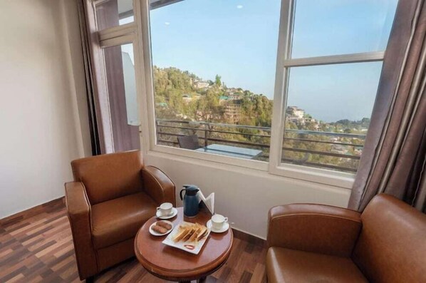 A room with two leather chairs, a small wooden table with breakfast items, and a large window offering a view of a hillside with buildings.