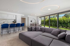 Ein modernes Wohnzimmer mit einer grauen Eckcouch, einem Essbereich mit blauen Stühlen und einer Küche mit weißen Schränken.