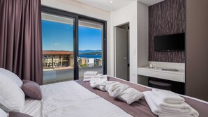 Een moderne hotelkamer met een bed, een badkamer met spiegel en televisie, en een balkon met uitzicht op zee.