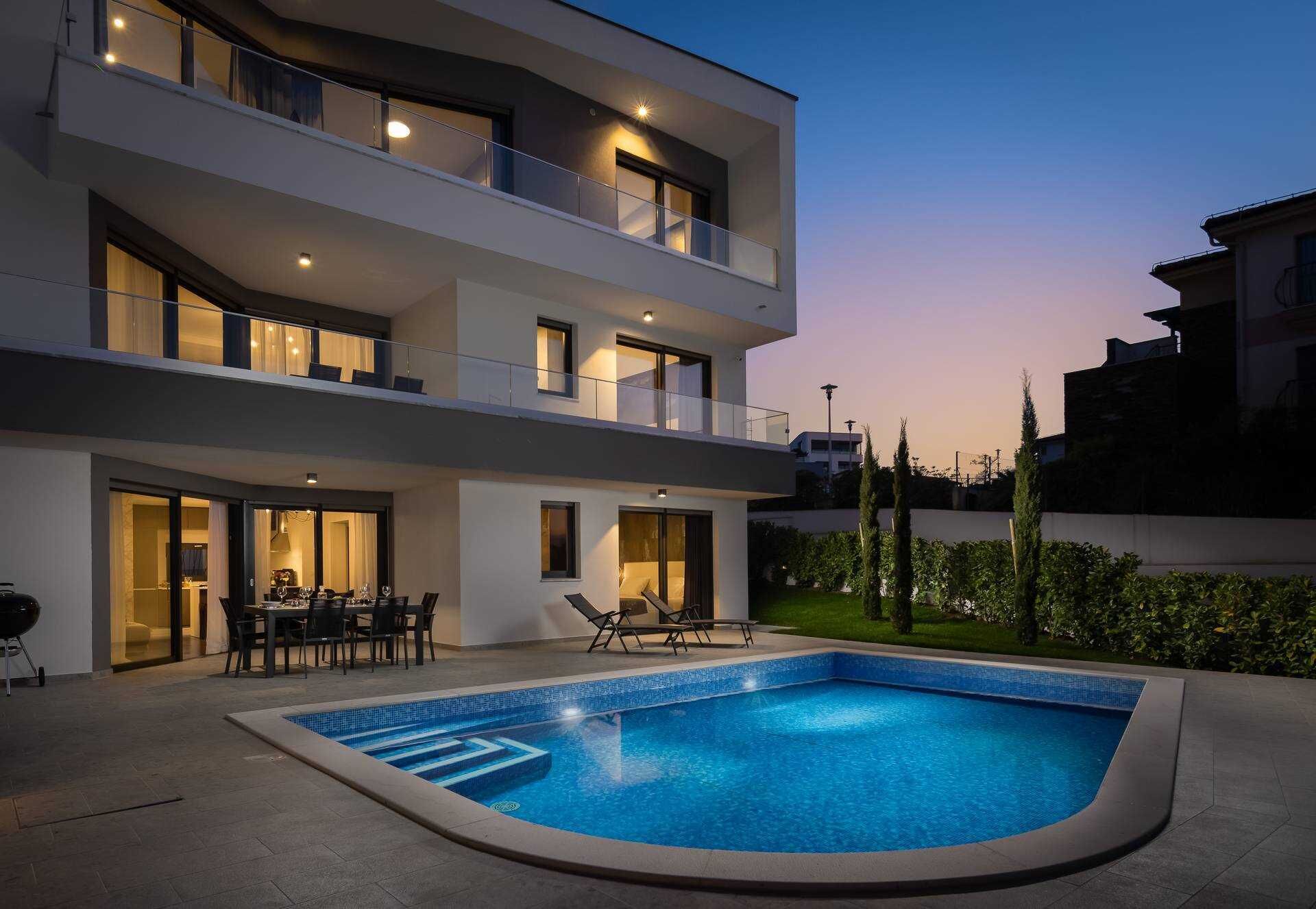 Een moderne, tweelaags woning met een zwembad, buitenmeubels en een verlicht balkon.