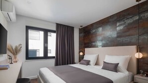 Ein modernes Schlafzimmer mit einem großen Bett, einem Schreibtisch mit Stuhl, einem Fenster mit Vorhängen und einem an der Wand montierten Fernseher.