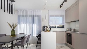 Eine moderne Küche mit Kochinsel, einem Essbereich inklusive Tisch und Stühlen sowie einem großen Fenster mit transparenten Vorhängen.