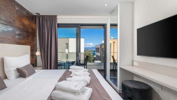 A modern hotel room with a large bed, a flat-screen TV, a balcony with a view of the city, and a neatly made bed with towels. - Villa Rova – Apartment with a Seaview (Malinska (Krk))