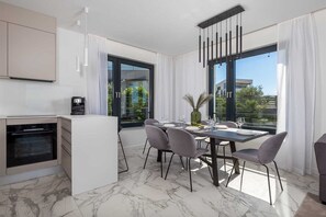A modern kitchen with a built-in oven, a dining area with a marble floor, and large windows offering a view of greenery and buildings. - Villa Rova – Deluxe apartment with balcony and beautiful sea view (Malinska (Krk))