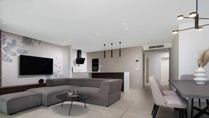 Ein modernes Wohnzimmer mit einem Ecksofa, einem Couchtisch, einem an der Wand montierten Fernseher und einem Essbereich mit Tisch und Stühlen.