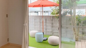 A room with a sliding glass door leading to an outdoor area with a red umbrella, two round white ottomans, and a bamboo fence. - Casa Estacion Hikone (Hikone)