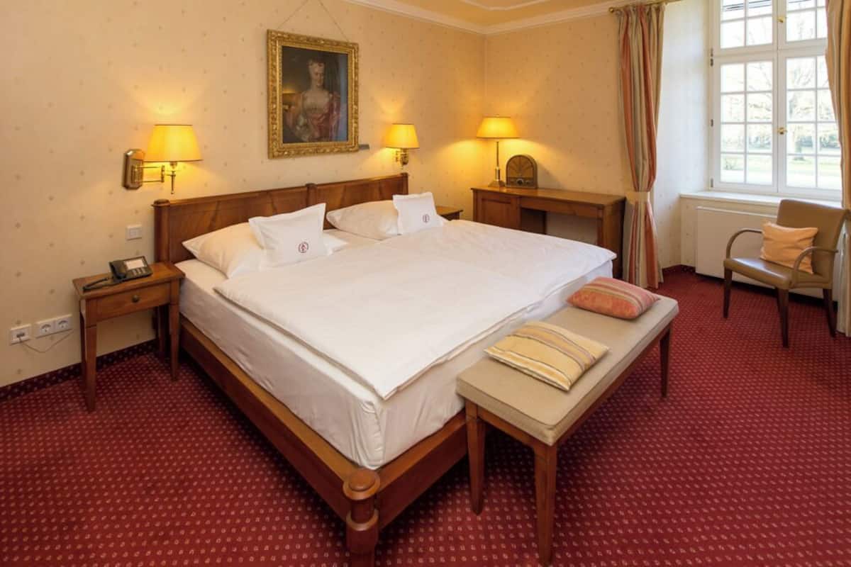 Ein Hotelzimmer mit einem großen Bett, zwei Nachttischlampen, einem kleinen Tisch, einem Sessel und einem Fenster mit Vorhängen.