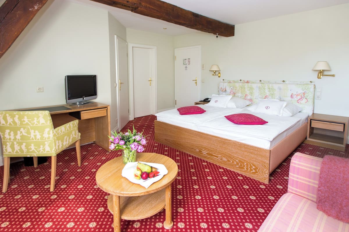 Ein Schlafzimmer mit einem Bett, einem Schreibtisch, einem Sessel, einem kleinen Tisch mit Obst und Blumen sowie einem Fernseher.