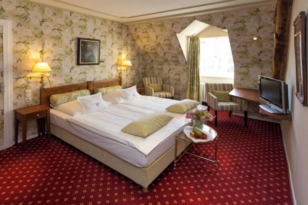 Ein Hotelzimmer mit einem großen Bett, zwei Nachttischen, einem Fernseher, einem Sessel und einem kleinen Tisch mit Blumen.