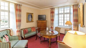 A room with a sofa, armchair, dining table, television, and a lamp. - Romantik Parkhotel Wasserburg Anholt (Isselburg)
