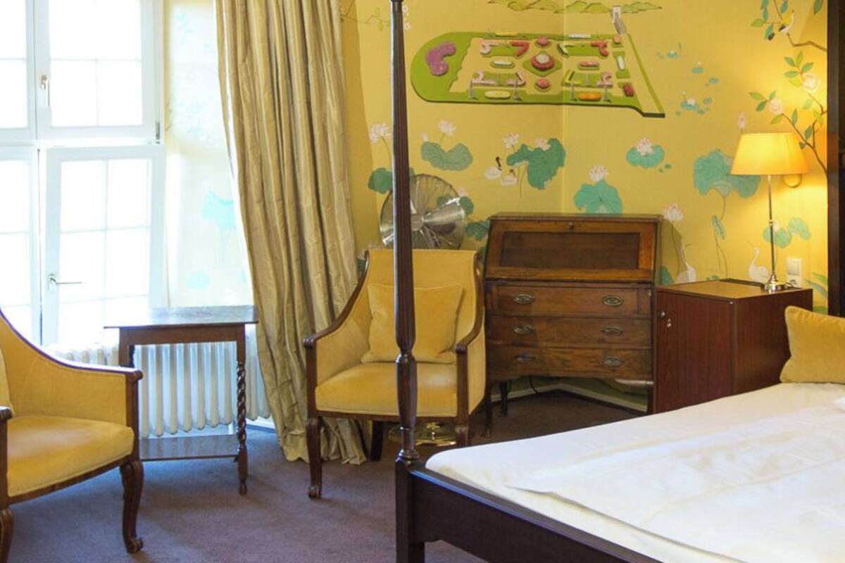 Ein Hotelzimmer mit einem Himmelbett, einem Nachttisch aus Holz, einem Sessel, einer Kommode und einem Fenster mit Vorhängen.