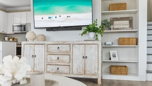 Una cocina con armarios blancos, un armario blanco con cajones, un televisor montado en la pared y una estantería con cestas de mimbre y un letrero que dice "beach house".