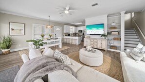 Una sala moderna con sofá, reposapiés y televisor de pantalla plana. Hay una cocina con armarios blancos y una mesa de comedor.