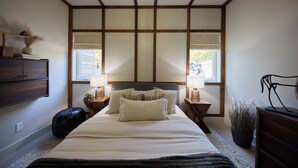 Quarto com cama grande, cabeceira de madeira, mesas de cabeceira com abajures, um vaso de planta e vista pela janela.