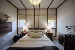 Une chambre à coucher avec un grand lit, une tête de lit en bois, des tables de chevet avec des lampes, une plante en pot et une vue par la fenêtre.