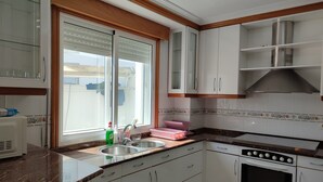 A kitchen with white cabinets, a stainless steel sink, a built-in oven, and a window with blinds. - Pensión Armonía (Caldas de Reis)