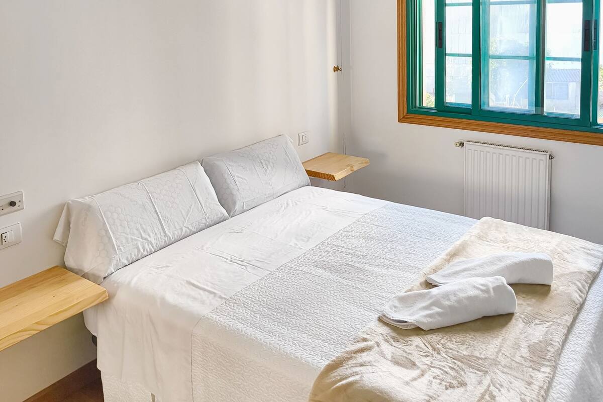 Un letto ben rifatto con piumone bianco, un comodino in legno, una piccola mensola in legno e una finestra con cornice verde.