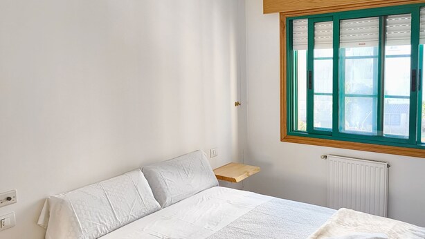 A bedroom with a bed, a window with blinds, a bedside table, and a radiator. - Pensión Armonía (Caldas de Reis)