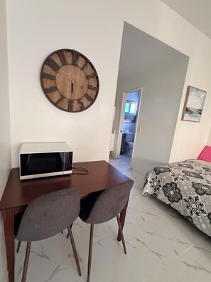 Een kamer met een houten wandklok, een witte magnetron, twee grijze stoelen, een bed met beddengoed met een patroon, en een deur naar een andere kamer.
