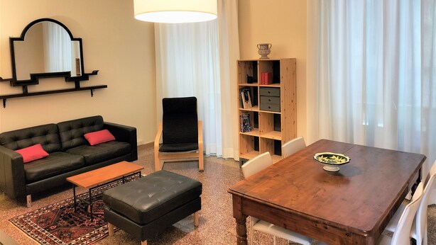 A modern living room with a black sofa, a wooden dining table, a black ottoman, a wooden bookshelf, and a large mirror on the wall. - Giannotti Vintage Apartment With Two Bedrooms (Firenze)