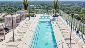 En pool på taket med kristallklart blått vatten, omgiven av solstolar och vilfåtöljer, med utsikt över stadens silhuett och grönområden.